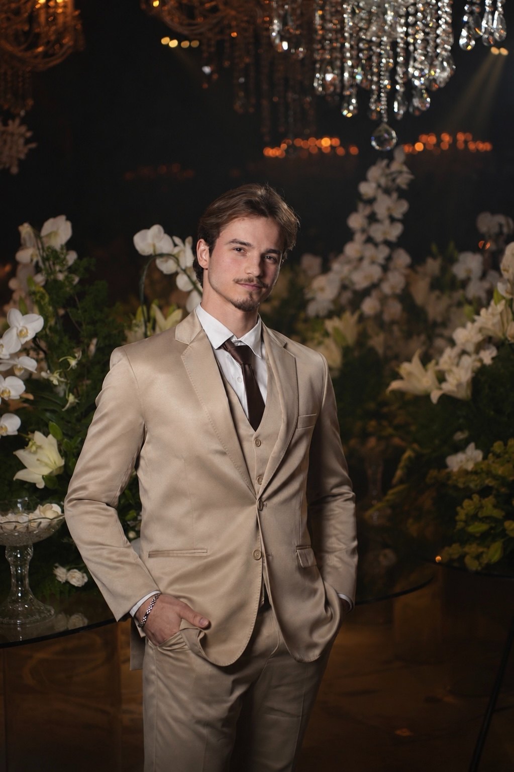 Igor de Castro de terno bege e gravata, em ambiente com flores e lustre